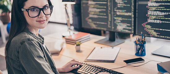 Eine junge Frau mit einer Brille sitzt an einem PC mit zwei Bildschirmen bei der Arbeit. Ihr Blick ist in die Kamera gerichtet.