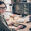Eine junge Frau mit einer Brille sitzt an einem PC mit zwei Bildschirmen bei der Arbeit. Ihr Blick ist in die Kamera gerichtet.