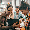 Drei Junge Leute lsitzen in einer Bibliohek an einem Tisch, auf dem Lernmaterialien liegen. Zwei junge Frauen schauen im Vordergrund auf ein Tablet und lächeln. Im Hintergrund schaut ein junger Mann zu, ebenfalls lernt. Links im Bild ist ein Bücherregal zu sehen.