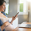 Schwangere sitzt mit Laptop am Schreibtisch und nutzt ein Smartphone.